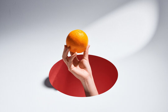 Woman's hand holding orange fruit on index finger through a hole cut in blue paper background