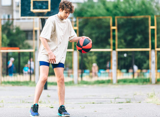 Cute teenager plays basketball at city playground. A boy holds basketball ball in his hands outside. Active life, hobby, sports for children