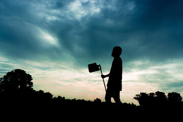 a silhouette of a person holding a long tool, standing in a field at sunset. There are trees and clouds in the background.