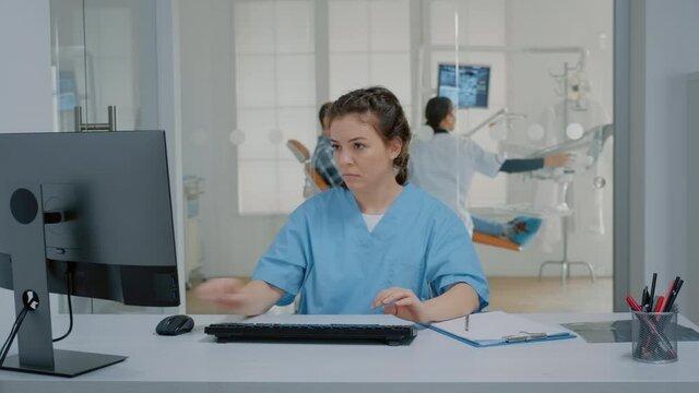 Dentistry nurse sitting at desk typing on computer keyboard working at dental clinic. Assistant with uniform using deivce while dentist examining teethcare of patient in background cabinet
