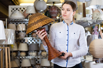 Cheerful positive smiling girl is choosing modern table lamp in furniture store..