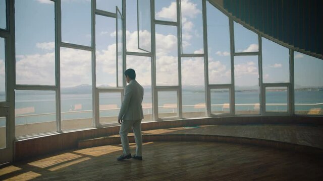 Businessman in a suit is walking towards a big window that looks at the sea . Back View of the Businessman wearing a Suit Standing in empty place . Shot on ARRI ALEXA cinema camera in slow motion .