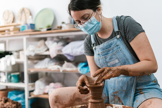 Female potter in glasses and mask working with clay - Powered by Adobe