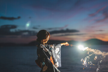 夕暮れの海で花火をする浴衣の女性