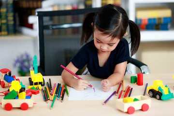 Small pretty pigtails hair preschooler kindergarten happy girl sit on chair drawing cartoon with colored pencils on paper on table full of plastic truck toys in living room at home in front bookshelf