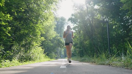 Slow motion sporty woman jogging on asphalt road among green trees. Tracking shot female athlete training in morning sunshine. Concept of fitness - Powered by Adobe