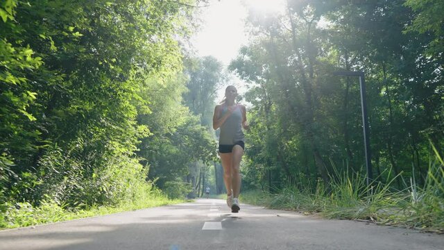 Slow motion sporty woman jogging on asphalt road among green trees. Tracking shot female athlete training in morning sunshine. Concept of fitness