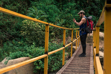 Fototapeta na wymiar young hiker man checking his phone in the middle of a bridge and carrying a red backpack surrounded by green bushes and trees in the rain forest on Ena hill in Costa Rica