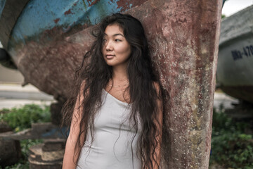 Portrait of beautiful young asian woman in white dress standing near the old ship and looking aside. Natural black curly long hair. Thinking face.