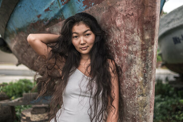 Portrait of beautiful young asian woman in white dress standing near the old ship and holding hair with hand. Natural black curly long hair. Looking at camera with copy space. Thinking face.