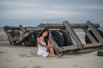 Charming young asian woman in white dress sitting on the beach and holding her leg. Brunette long curly hair. Romantic photo.
