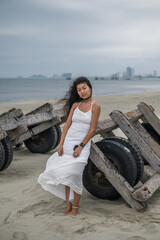 Charming young asian woman in white dress sitting on the beach and looking at camera with copy space. Brunette long curly hair. Romantic photo.