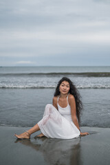 Charming young mongol woman in white dress sitting on the beach. Black long curly hair. Looking at camera with copy space. Romantic photo. Ocean view.