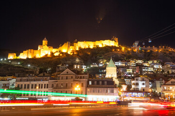Night city of Tbilisi. In the background is a cascade of buildings with architecture typical of the region. Narikala Fortress is illuminated by the initial lighting. In the foreground are long exposur