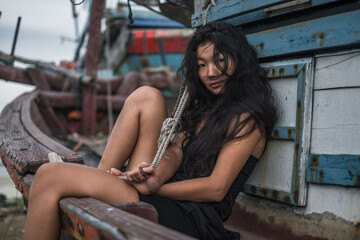 Naklejka na ściany i meble Portrait of charming young asian woman sitting on the old wooden ship. Street style portrait. Black long curly hair. Looking at camera with copy space. Wind in her hair. 