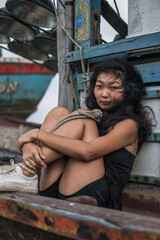 Portrait of beautiful young asian woman sitting on the old wooden ship. Street style portrait. Black long curly hair. Wind in her hair. 