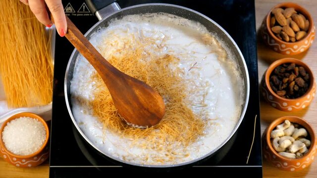 Female hand mixing the Semiya into a pan of boiling milk with a wooden spatula. Sevai  sugar  cashew nuts  and raisins kept in earthen pots - Indian dessert for Eid festival