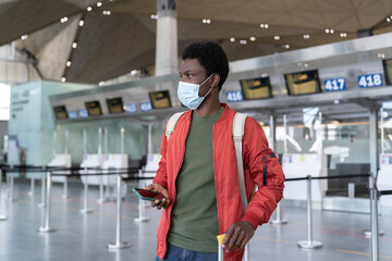 Fototapeta premium Young black male traveler in airport wait for flight use smartphone wear mask in covid-19 epidemic. African man passenger chat online before departure. Safe travel and coronavirus new reality concept