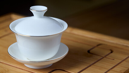 Gaiwan on chaban selective focus. Tea ceremony gong fu cha. Chinese white pottery Dehua white Chinese porcelain gaiwan bowl tea cup and lid plate with gold border on bamboo teaboard tea tray chaban