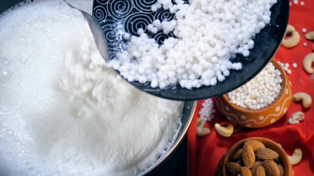 Preparation of Indian dessert - Sabudana Kheer in an Indian home during festival. Female hand adding white tapioca pearls with a spoon into the boiling milk to make delicious Kheer at home