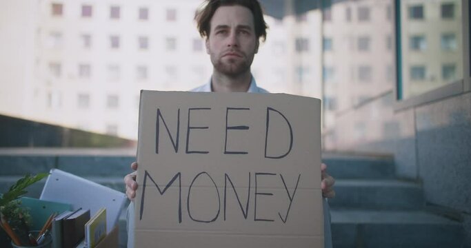 Zoom in portrait of young workless manager sitting outdoor with poster Need Money, looking for new job, slow motion