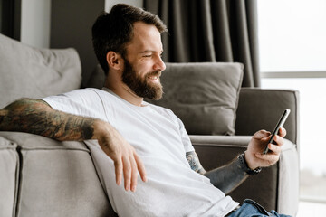 Bearded european man using mobile phone while sitting on floor