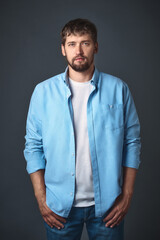 This young confident man with a beard in stylish casual clothes poses in the studio against a gray background. Portrait of a handsome guy of Caucasian appearance.
