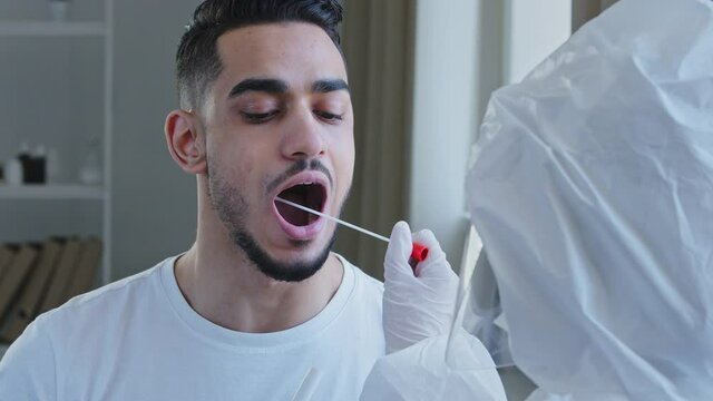 Arabic young handsome man Spaniard sick male patient ethnic guy opens mouth for pcr analysis covid-19 virus test with unrecognizable doctor nurse medical worker using equipment cotton swab in lab
