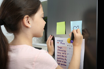 Little girl drawing heart on note near to do list in kitchen, closeup