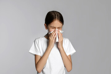 Little girl blowing nose into paper tissue on light grey background. Seasonal allergy