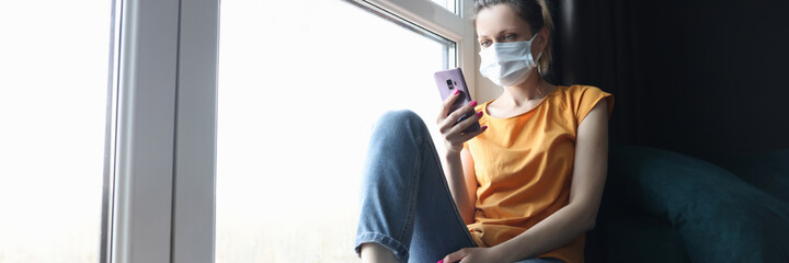 Fototapeta na wymiar Woman in protective medical mask sitting on windowsill with mobile phone in her hands