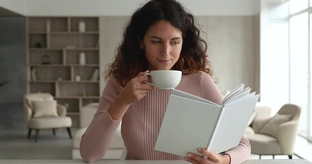 Positive happy attractive millennial hispanic latin woman involved in reading favorite novel literature in paper book, drinking hot coffee or tea, enjoying carefree leisure weekend time alone at home.