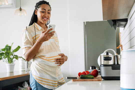 Black pregnant woman smiling and using mobile phone in kitchen - Powered by Adobe