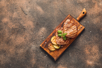 Wooden board with tasty beef steaks and grilled vegetables on grunge background
