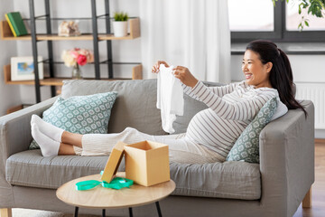 pregnancy, holidays and people concept - happy smiling pregnant asian woman opening gift box with baby's bodysuit at home