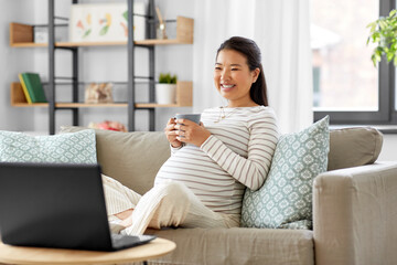 pregnancy, rest, people and expectation concept - happy smiling pregnant asian woman with laptop computer sitting on sofa at home and drinking tea