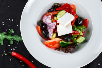 greek salad with olives, bell peppers, onions and feta cheese in a white plate on a dark background with ingredients close-up top view