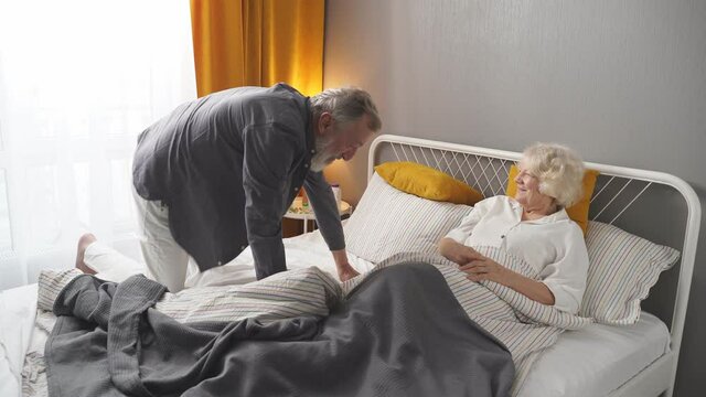 Positive caucasian elderly couple have fun. Laugh while lying on bed at home, in domestic clothes, morning, daily routine of retired people, harmonious relationships.