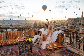 A couple and their romantic breakfast in Cappadocia on the amazing background of hundred flying balloons. - obrazy, fototapety, plakaty