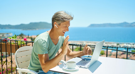 Joyful man wearing headphones communicating with friends via video computer call during sea resort summer vacation