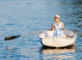 Foto op Canvas An elegant blonde woman in a hat sits on a boat and enjoys the tranquil lake during her summer vacation. © VIK