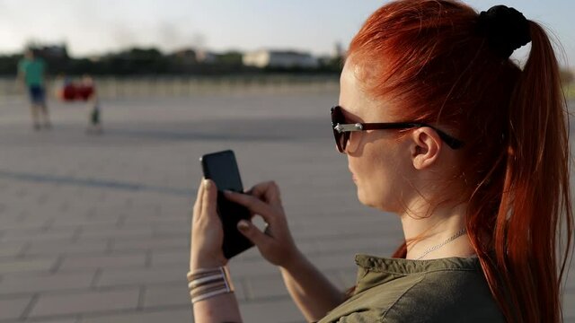 a red-haired woman in sunglasses with a phone in her hand in the park, modern communication technology