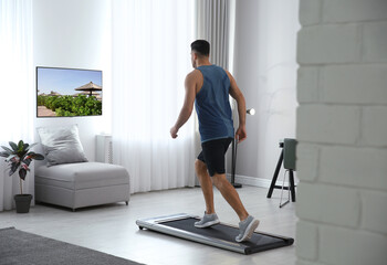 Sporty man training on walking treadmill and watching TV at home - obrazy, fototapety, plakaty