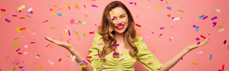 cheerful woman with outstretched hands near confetti on pink, banner