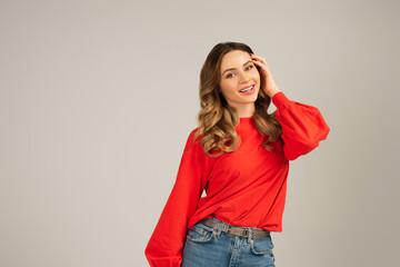 happy woman in sweatshirt looking at camera and adjusting curly hair isolated on grey