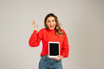 amazed woman in wireless earphones holding digital tablet with blank screen isolated on grey