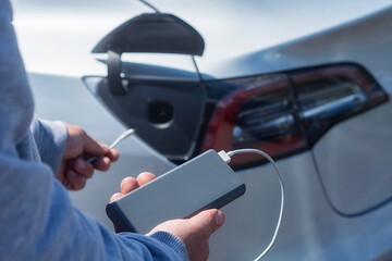Electric vehicle charging. Man holds a power bank with a cable in his hands against the backdrop of an electric car.