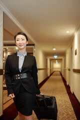 Contetn attractive young Asian woman in formalwear being on business trip walking with luggage along hotel corridor