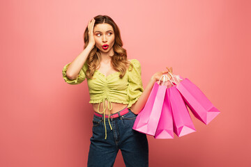 shocked woman holding shopping bags and pouting lips isolated on pink