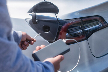Electric vehicle charging. Man holds a power bank with a cable in his hands against the backdrop of an electric car.
